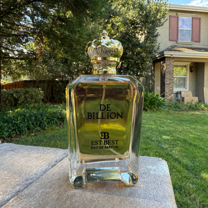 DE BILLION perfume bottle photographed outdoors in natural light, highlighting the clear glass and gold accents.
